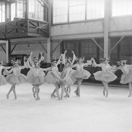 A black and white photograph of ten tutu-costumed skaters in various poses, many standing up on toe picks with arms overhead in ballet poses. The group is in an indoor ice rink with large windows.