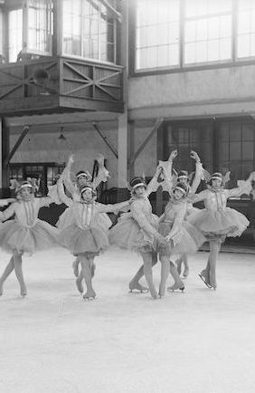 A black and white photograph of ten tutu-costumed skaters in various poses, many standing up on toe picks with arms overhead in ballet poses. The group is in an indoor ice rink with large windows.