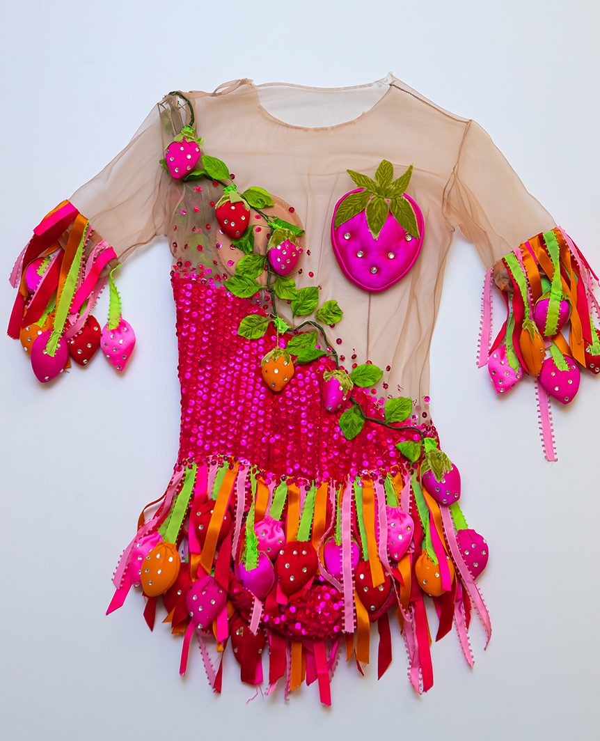 Skating dress lying flat on a white background. The bodice is beige sheer fabric from the neck to waist, including the top of the arms. A strawberry vine obscures the breast area. Ribbons create the skating skirt and fringe at the above-the-elbow sleeve.