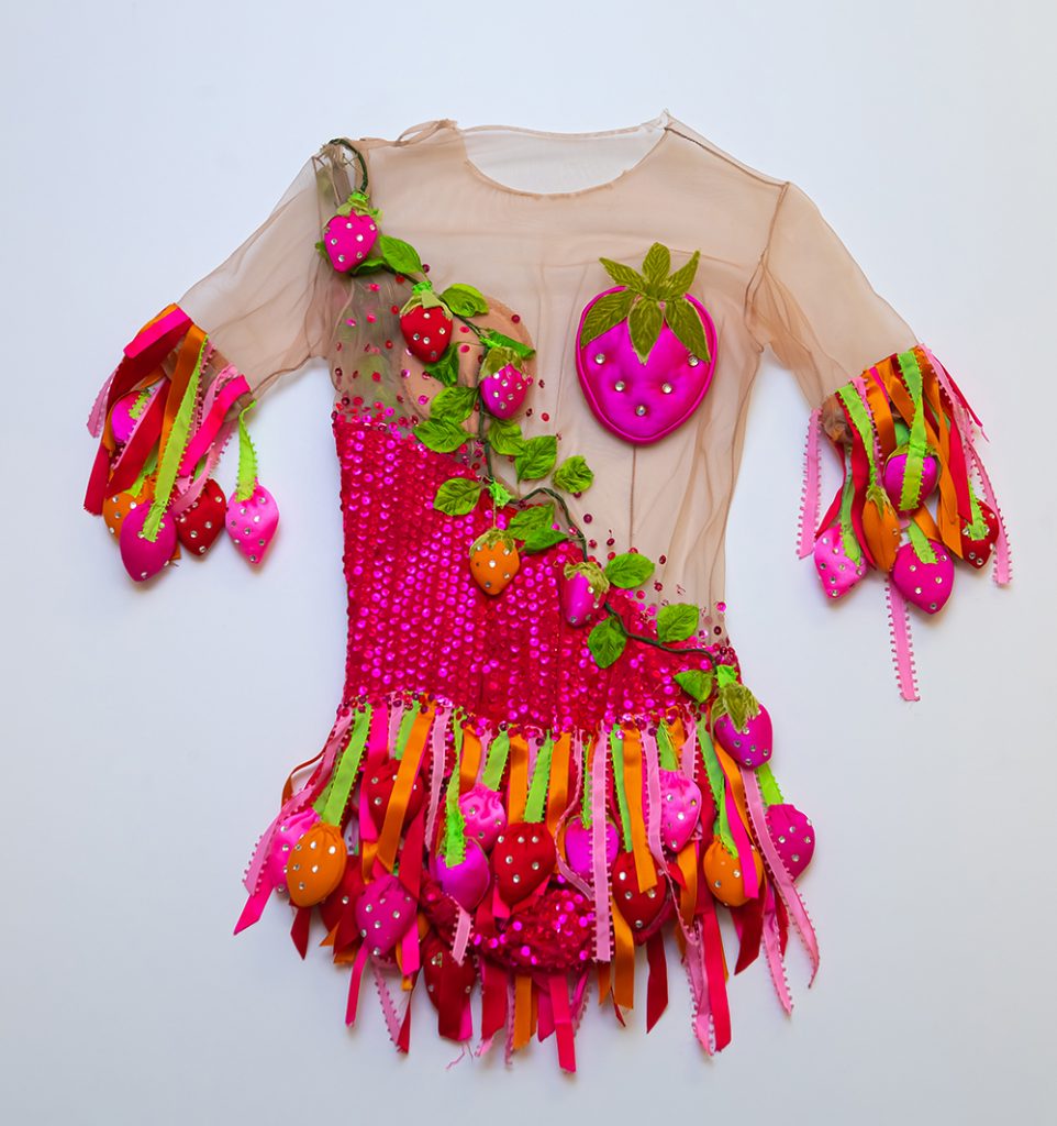 Robe de patinage posée à plat sur un fond blanc. Le corsage est fait d’un tissu beige transparent du cou à la taille, y compris le haut des bras. Une grappe de fraises masque la poitrine. Des rubans créent la jupe et les franges au niveau de la manche, plus précisément au-dessus du coude.