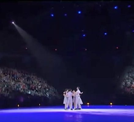 The seven skaters together at centre ice. The spotlights create a blue hue on the ice with the skaters highlighted in a yellow hue.