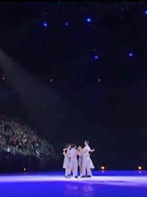 The seven skaters together at centre ice. The spotlights create a blue hue on the ice with the skaters highlighted in a yellow hue.