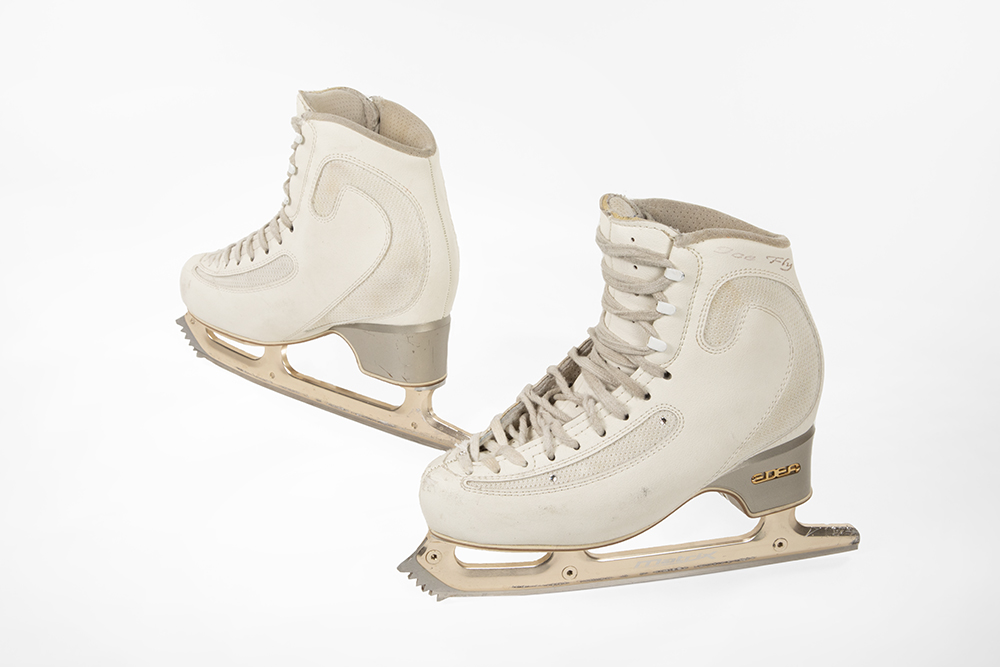 A pair of small size white skating boots have a swirl inset around the ankle area. “Ice Fly” is written the upper edge of the ankle. Gold metal letters “EDEA” are mounted in the stacked leather heel.