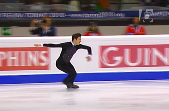 Patrick Chan, in a black jacket and pants costume with white trim at the neck and sleeve cuffs, performs a routine filled with complex footwork, jumps and a travelling jump sit spin variation. An audience watches and applauds in the stands.
