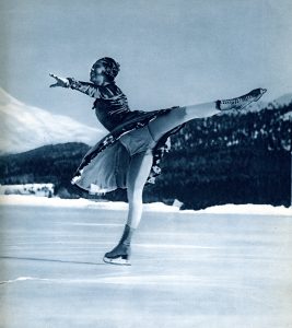 A black and white photo of a woman, Sonja Henie, skating on an outdoor arena with left arm and left leg extended in the bent knee style. She wears a knee length skating dress. Trees and a snowy mountains are seen in the distance.