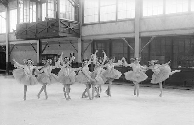 A black and white photograph of ten tutu-costumed skaters in various poses, many standing up on toe picks with arms overhead in ballet poses. The group is in an indoor ice rink with large windows.