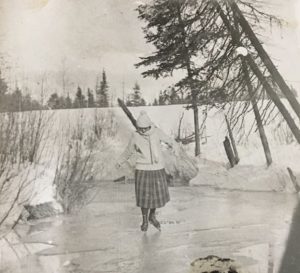 A black and white photo of a woman in a toque, heavy sweater and mid-calf length tartan skirt, skating on a frozen pond.