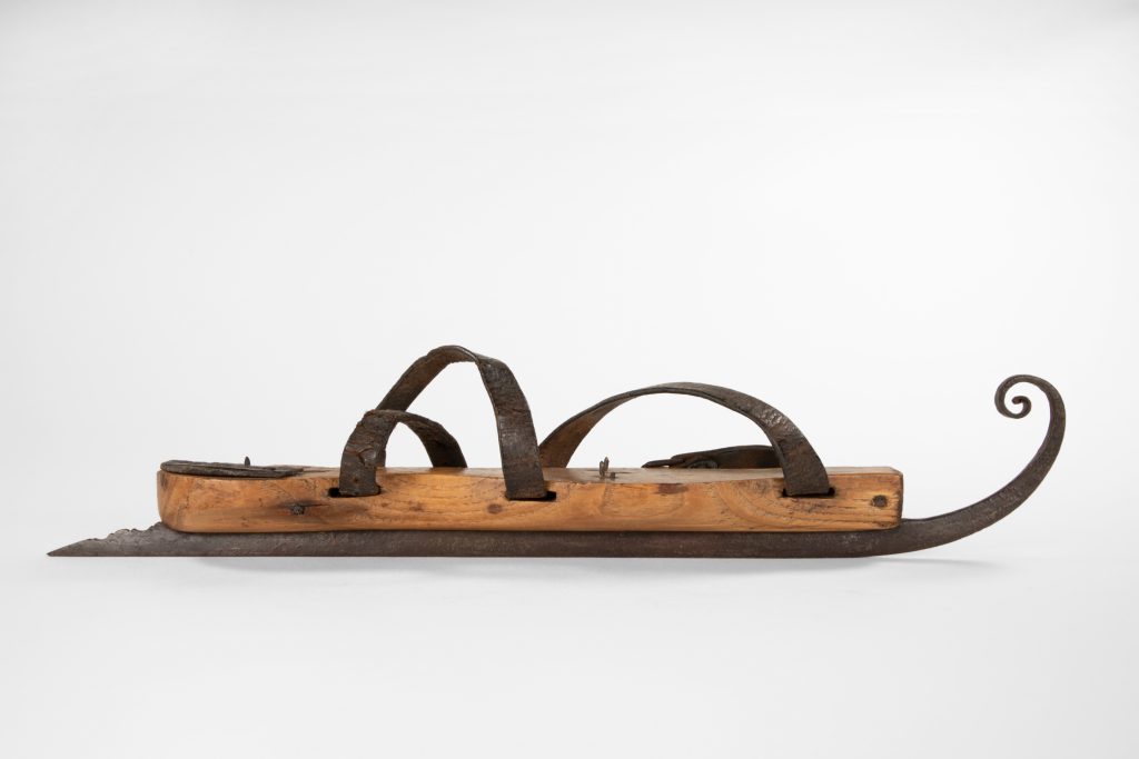 A single skate blade with a very tightly curled toe tip is mounted onto a wooden platform for the foot. The heel extension of the blade is decoratively carved. Shown in profile view.