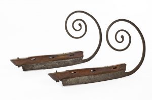 Pair of skate blades in profile view. At the front, the blade extends up and over the wearer’s toe, curling like a snail’s shell.