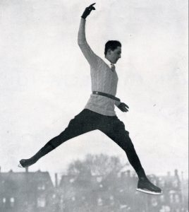 A black and white photo of Bud Wilson mid-air in a jump with legs is a split ‘V’. He wears a belted knit sweater, dark jodhpurs, dark skates and dark gloves.
