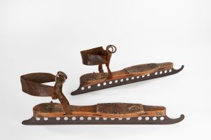 Pair of skates designed with 16 small holes along the length of each, and a slight up-curl at the toe. The blades strap-on to the wearer’s foot by way of the leather straps at heel. Missing leather straps at the forefoot.