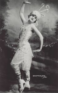 A black and white photo of a woman in a diaphanous beaded costume, posing in skates.