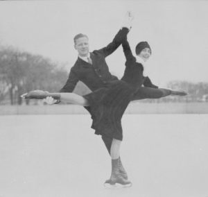 A black and white photo of a man and woman posing, each with one leg extended, joining hands, and touching the other’s extended foot.