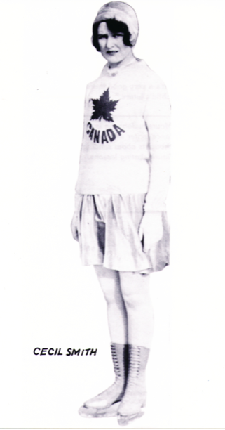 A black and white photo of Smith in skates and the team ‘Canada’ sweater, looking toward the viewer. Her light coloured skates are cut high on the lower leg, ending below her calf muscle.