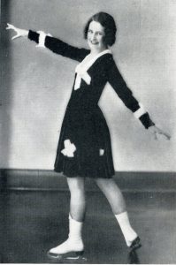 A black and white photo of a smiling woman in a knee length black dress with white trim and white skates.