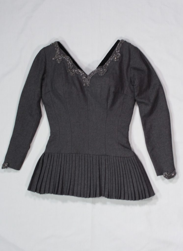 A photo of a short, fitted, dark wool one-piece skating dress with long sleeves and many small pleats on the full skirt. The V-neck is embellished with beaded trim.