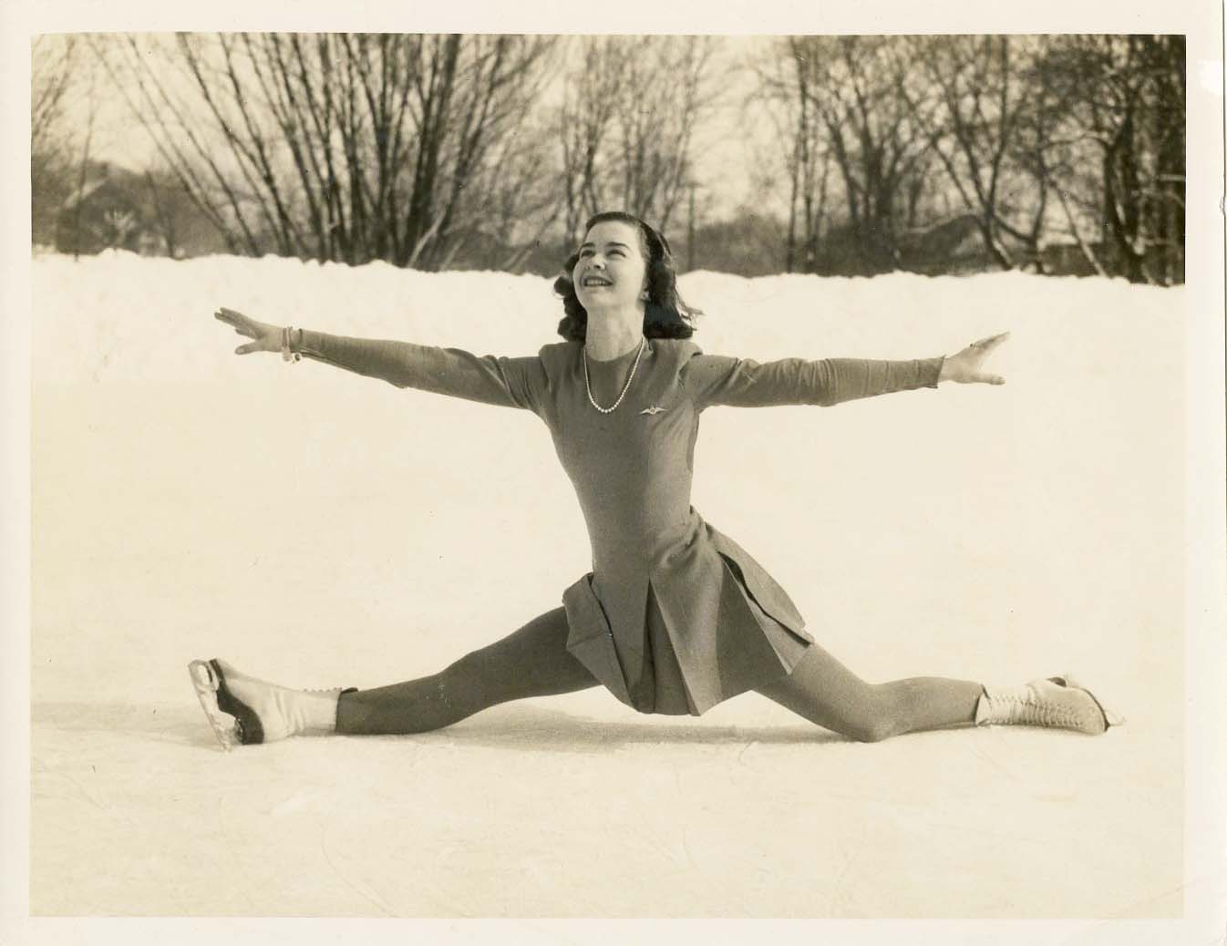 Une photo en noir et blanc de Barbara Ann Scott exécutant un grand écart sur une patinoire extérieure.