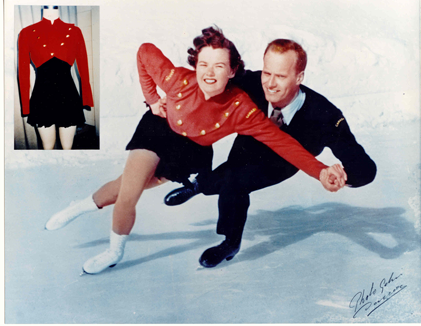 A colour photo of a Bowden and Dafoe pair skating towards the viewer. Bowden wears a dark suit, blue shirt and necktie, while Dafoe wears a red bolero length jacket and black skating dress. 