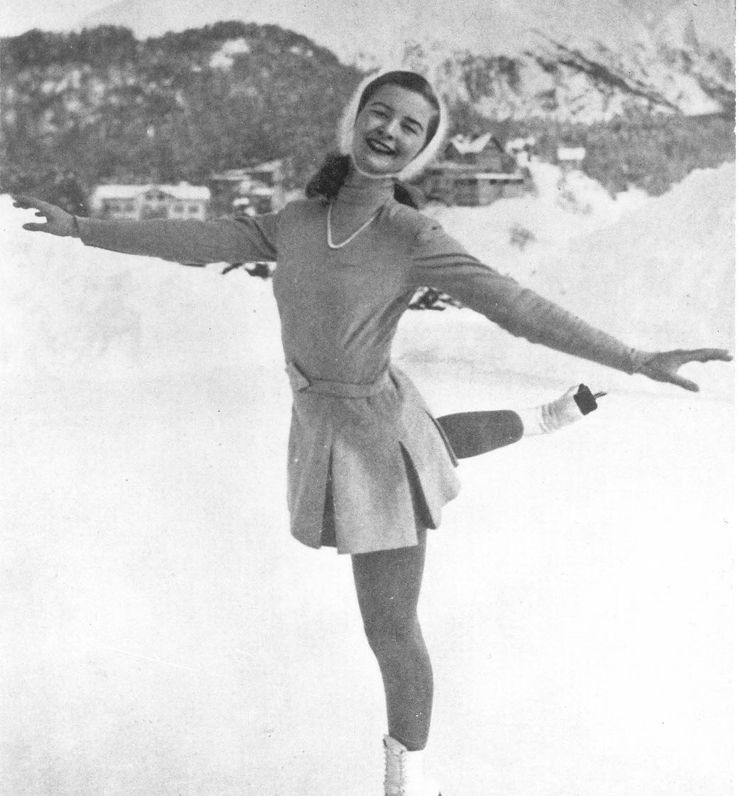 Une photo noir et blanc à partir de la taille d’une femme souriante, sur une patinoire extérieure avec des bâtiments bas et des montagnes en toile de fond.