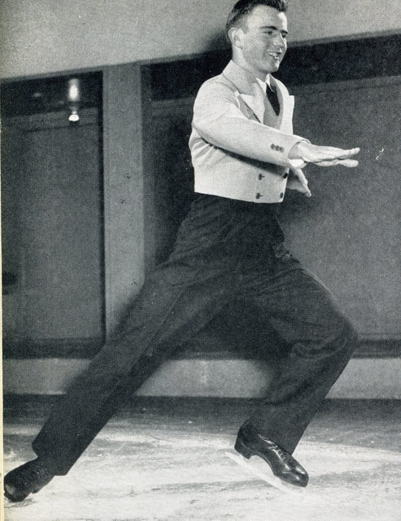 Photo en noir et blanc d’un homme patinant dans une patinoire intérieure vers le côté droit de l’image. Sa jambe gauche est légèrement pliée tandis que sa jambe droite est étendue derrière lui sur la glace. Il porte une courte veste croisée ajustée et un pantalon de laine noir à plis, ainsi que des patins noirs.