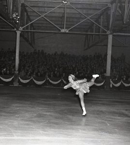 Photo en noir et blanc de Barbara Ann Scott en costume de patinage scintillant sur une patinoire intérieure. Elle patine en spirale avec sa jambe gauche étendue vers l’arrière pendant qu’un public assis regarde en arrière-plan.