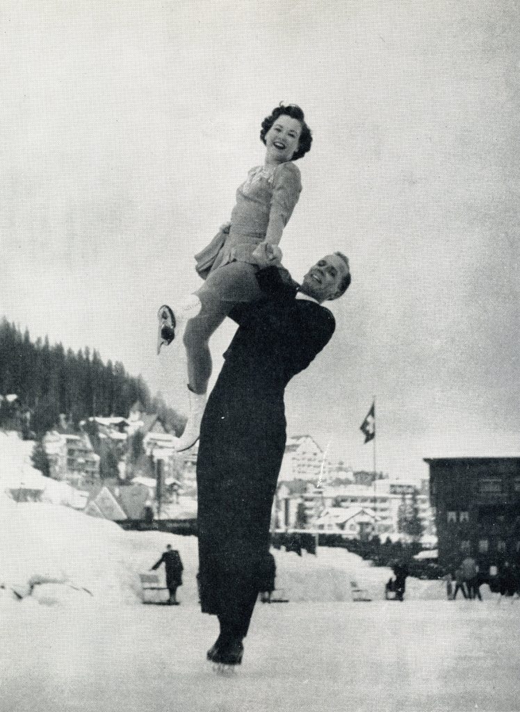 Photo en noir et blanc de Norris Bowden soulevant Frances Defoe au-dessus de sa tête tout en patinant, sur une patinoire extérieure. D’autres patineurs, un long bâtiment et des montagnes sont en arrière-plan.
