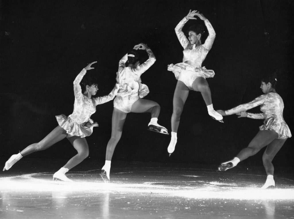 A black and white photo of a young woman preforming a skating jump on a darkened ice rink. 