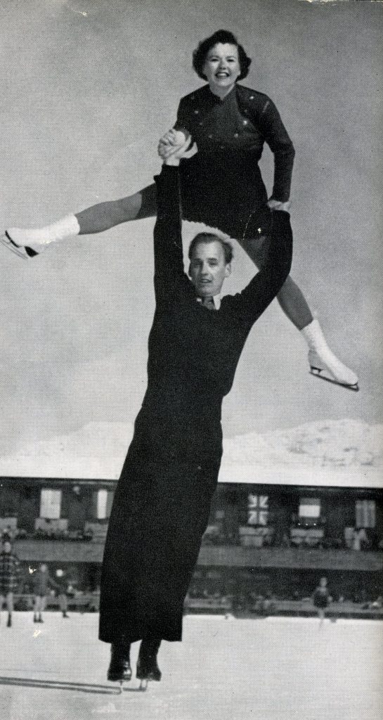 A black and white photo of Norris Bowden lifting Frances Defoe above his head while he skates on an outdoor rink. Other skaters, a long building and mountains in the background.