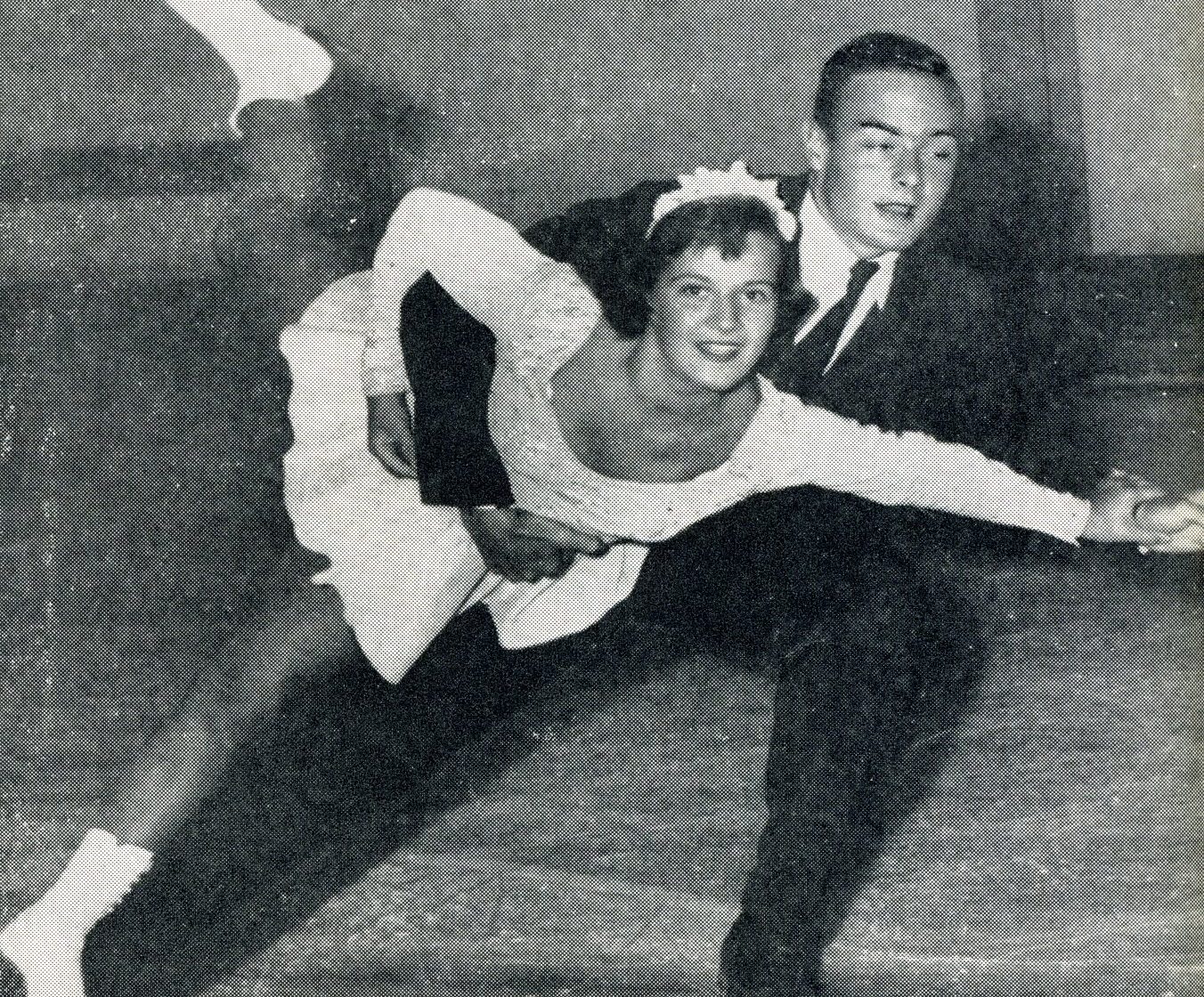 Photo en noir et blanc d’un homme et d’une femme qui patinent ensemble en direction des spectateurs sur une patinoire intérieure. Il la tient fermement à la taille. La femme sourit et porte un costume blanc à manches longues. L’homme porte un veston, une chemise blanche et une cravate foncée.