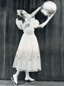 A black and white photo of a costumed female skater posing with a large silver sphere.