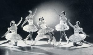 A black and white photo of five costumed female skaters posing for the camera. The background is spot-lit and the central skater is perched on a white crescent, which might be a moon beam.