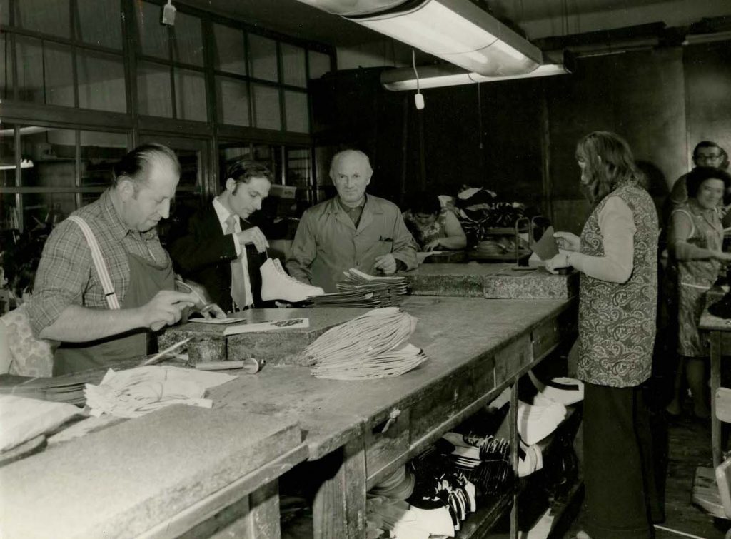 Don Jackson in suit and tie holds a white skate boot while three boot makers continue work around the large waist-height bench. Fluorescent light overhead.