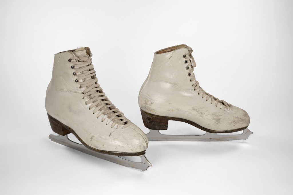 Pair of white custom made skates with four speed lacing hooks up the ankle. Three-quarter view showing the outer side of one skate, the inner side of the left skate.