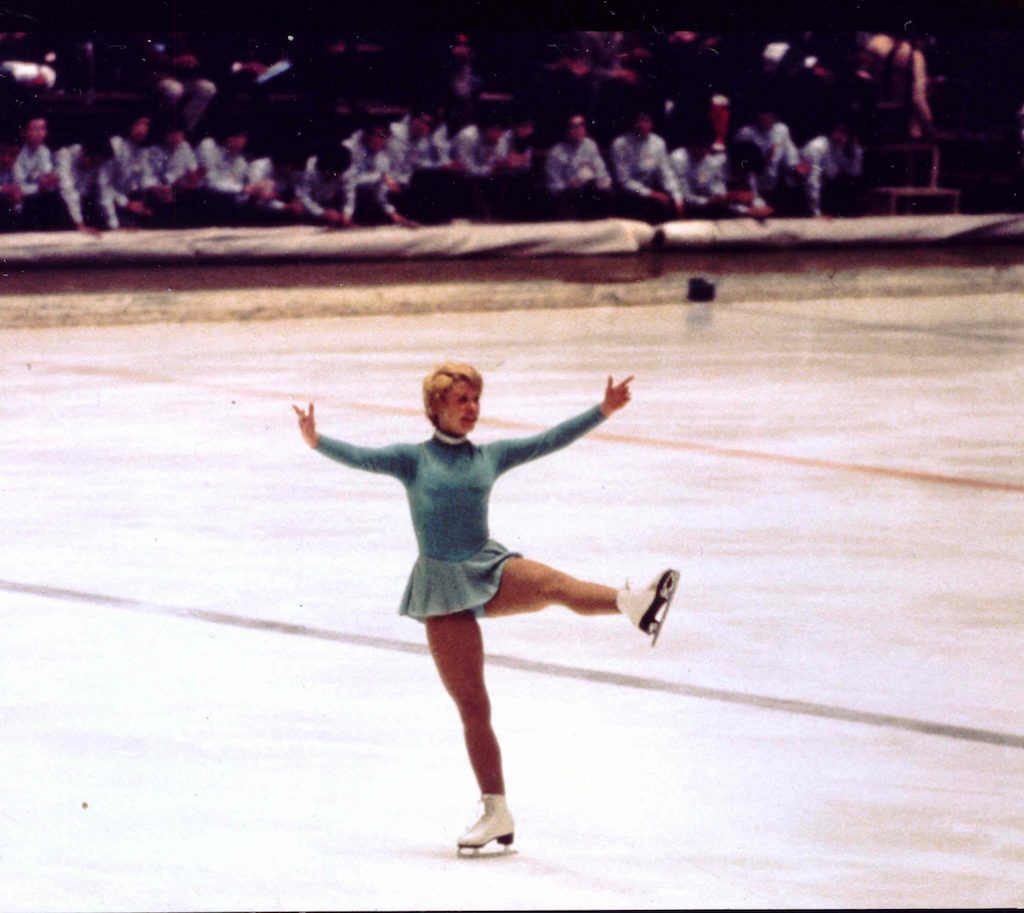 Karen Magnussen se prépare pour faire une révérence en déplaçant la jambe gauche d’avant en arrière en demi-cercle. Elle porte une robe de patinage vert pâle.