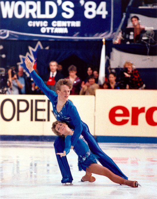 Martini holds Underhill with one hand as she does the splits between his legs. They are wearing royal blue glossy skating outfits. He in long pants and she in short skating leotard with skirt. Her skates are beige.