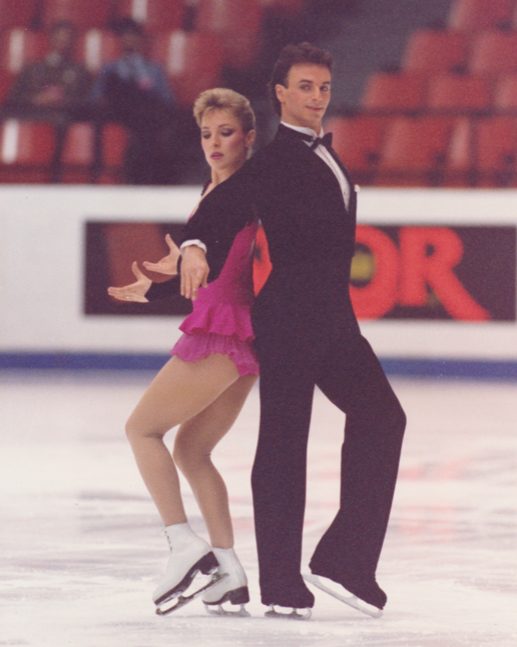 Standing back-to-back and stationary, Wilson and McCall interpret the music with arm movement and facial expression. She in a short pink skating dress; he in black with white shirt showing at neck and sleeve cuff. Bow tie.