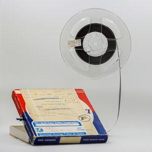 Photo of a reel to reel magnetic tape in a clear plastic reel. The reel’s box is on the bottom left with the lid propped open. Visible text reads: “Feb. 75, full track, Ampex recorder, Lynn Nightingale Minto Figure Skating Club.” Sticker lower down reads: “1977 World Figure Skating Championships”.