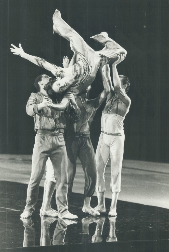 Black and white photo of three male dancers, wearing warm-up outfits, holding an inverted Toller Cranston above their heads. Toller has skates on and his feet are at the top of the photo. His dramatic facial expression and arm position are at the centre of the photo. He wears a stretch jumpsuit with sequins across the chest. The dancers wear dance slippers and stand on a mirror-finish floor.