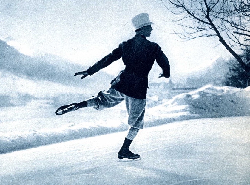 A black and white photo of a man, Grafström, skating away from the viewer with his left leg extended behind him. His arms are gracefully out for balance at hip level. He wears a white hat, knickerbockers, thick knee high socks and dark skates on an outdoor rink. 