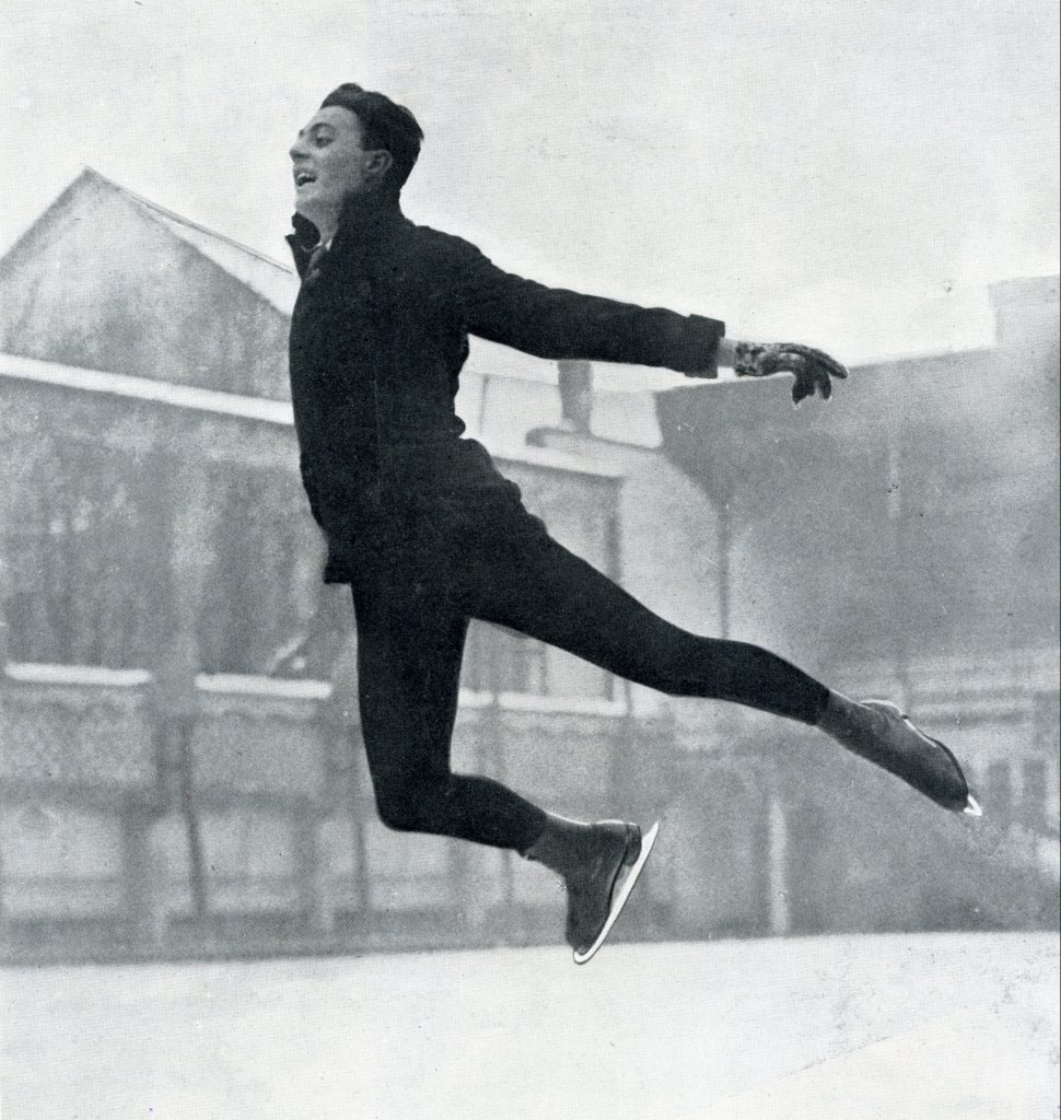 A black and white photo of a smiling male skater leaping forward with arched back and both arms and legs extended slightly behind him. He wears a narrow-cut jacket and tie with fitted black tights.
