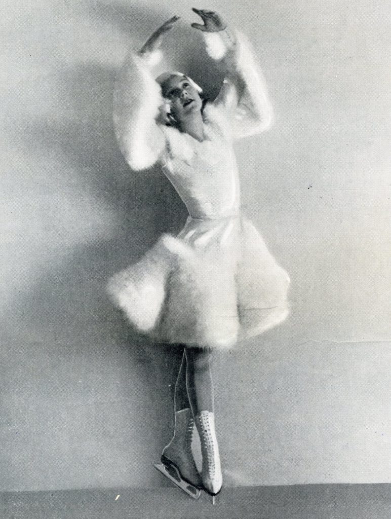 A black and white photo of Sonja Henie in tall white skates above the ankle and a fancy white skating costume with fur or feather sleeves and skirt. She is posing on pointed toes with hands above her head in a ballet position. 