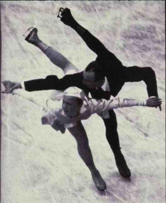 Photo en plongée en noir et blanc d’un homme et d’une femme patinant vers le spectateur, la jambe gauche étendue, avec le genou plié. 