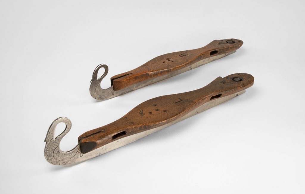 Pair of embellished skate blades with wood foot mounts. No leather straps remain.