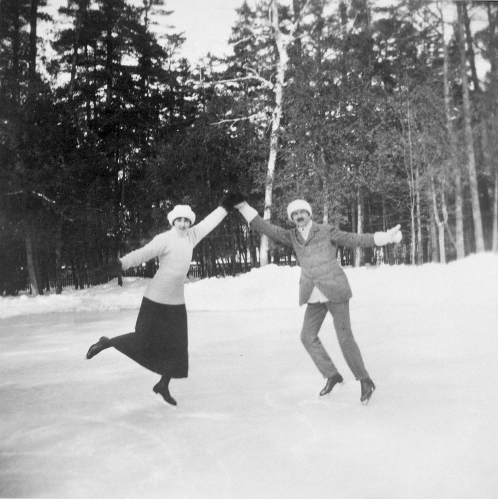 Une photo en noir et blanc d’un homme et d’une femme habillés de vêtements d’hiver; ils se tiennent sur la pointe de leurs patins et se donnent la main en posant pour la caméra.