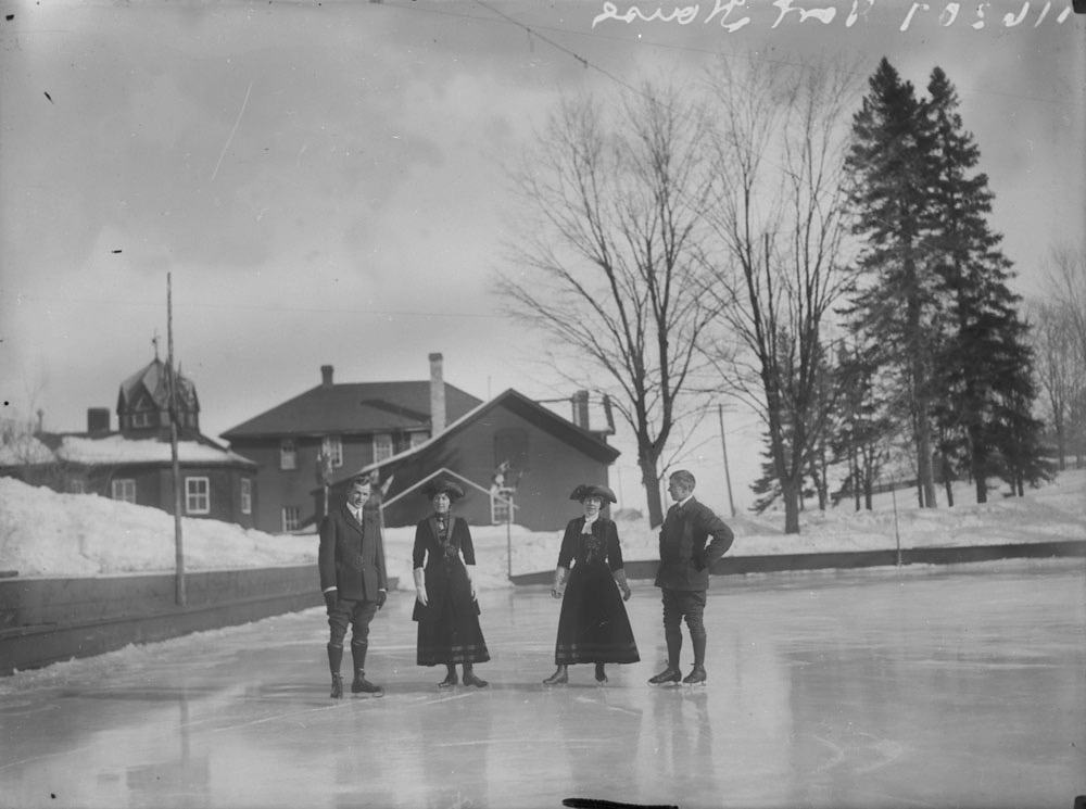 Black and white photo of two women in long skirts and two men standing on skates at a skating rink with two buildings behind them.
