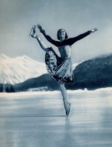 A black and white image of a smiling Sonja Henie skating outdoors with both arms and right leg extended upwards. Her skating boots end quite high on the leg, above the ankle. She is on a frozen body of water with trees and mountains in the distance.