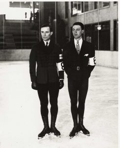 A black and white photo of two men in dark, tight-fitting suits and neck ties, facing the camera in an indoor rink. White arm bands with an identifying number on their left arm at elbow height. Their skates are cut high on their lower legs. Grafström’s skates are highest; cut just under his calf muscle.