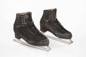 Photo of a well-used pair of black leather skates, laced mid-way up, standing in a parallel position.