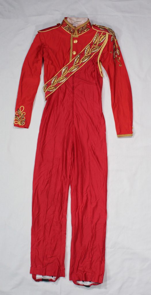 A red one-piece military-inspired men’s costume. Standup collar and epaulets have gold trim around the edges and sequined patterns. A diagonal sash across the chest. Red strapping at bottom of the leg to attach under the skating boot.