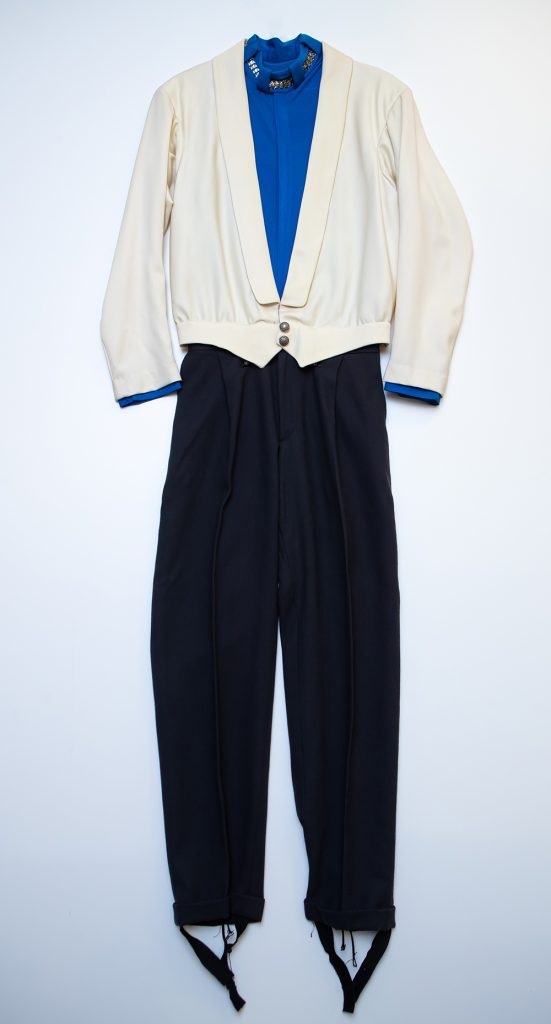 An off-white dinner jacket styled top with two round metal buttons at waist. Royal blue shirt under the jacket has a mock-turtle neck collar. The black, pleated pants have baggy legs. The bottom of the pant legs each has a black strap to attach underneath the skating boot.